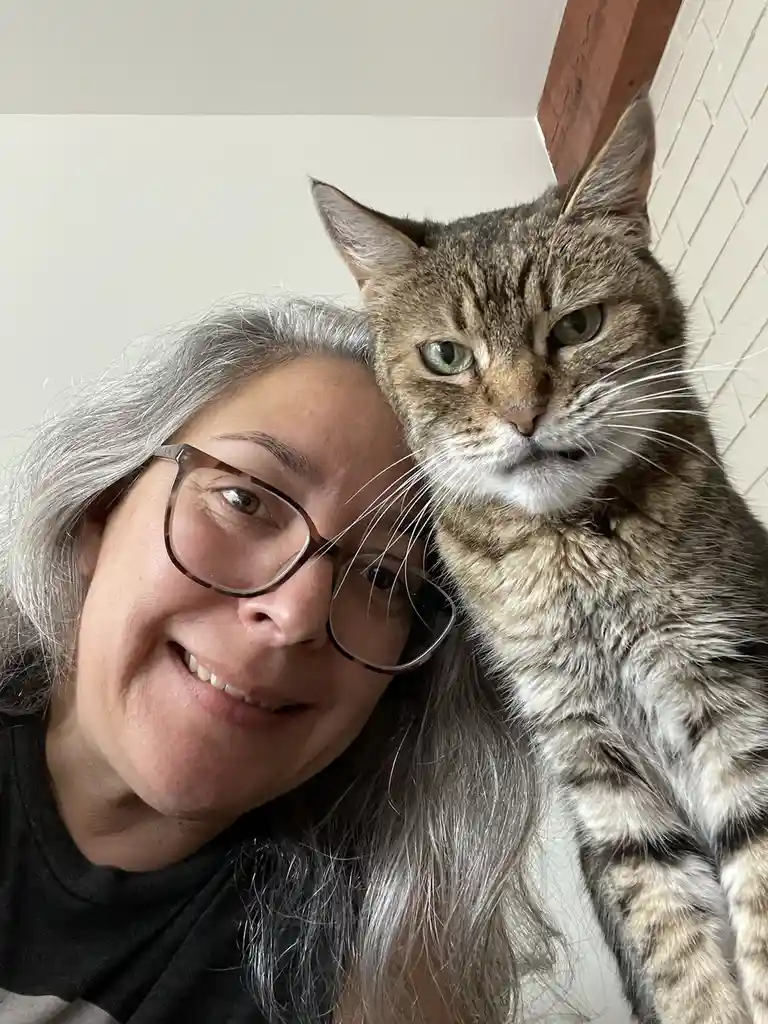 A woman with gray hair and glasses smiles at the camera while a tabby cat with green eyes rests its head on her shoulder…