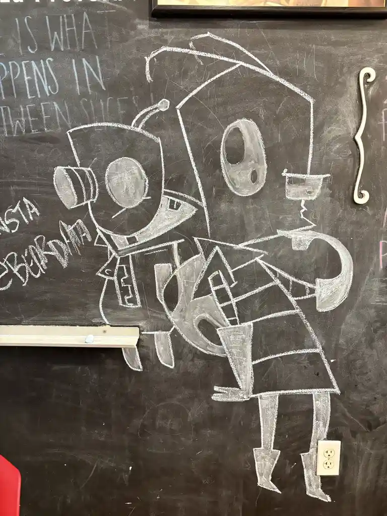 A chalk drawing on a blackboard depicts two cartoonish robot-like characters in a romantic embrace, one with a top hat and…