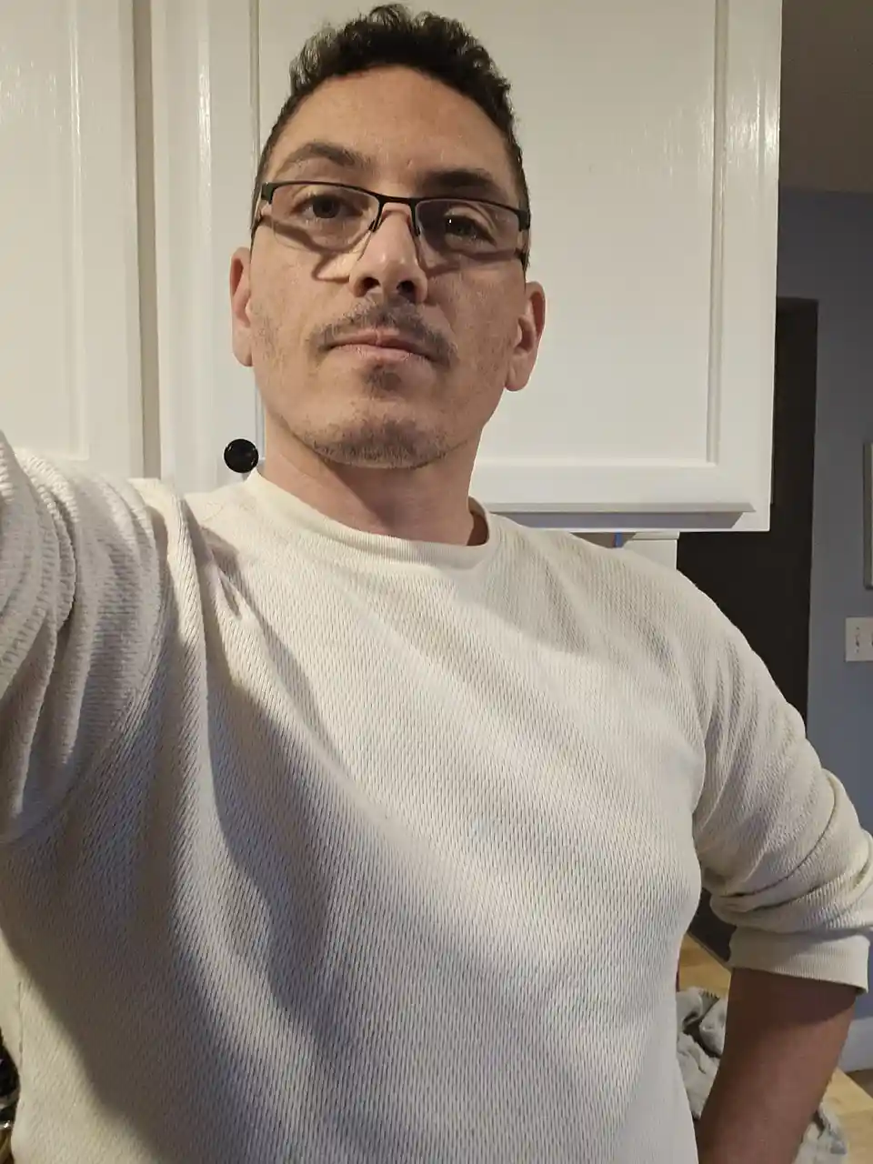 A man with dark hair, glasses, and a light-colored ribbed sweater takes a selfie in front of white kitchen cabinets. He has…