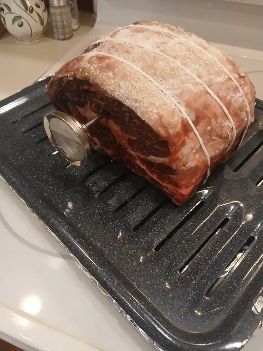 A large raw beef roast, tied with twine and covered in coarse salt, sits on a black metal roasting rack. A meat thermometer…