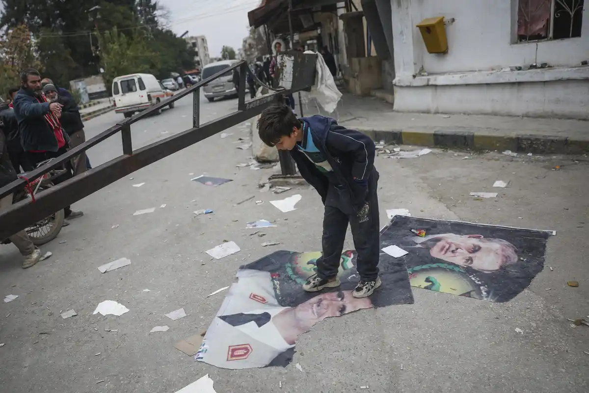 A young boy stands on a torn political poster featuring a man's face, looking down at it with his hands in his pockets. The…