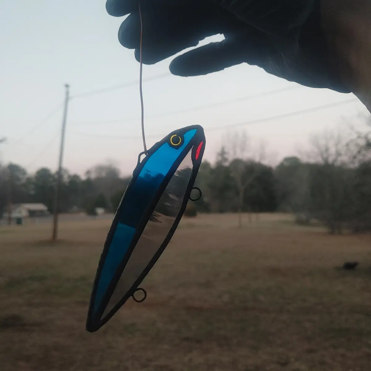 A hand in a black glove holds a stained glass fishing lure shaped like a blue and silver fish, suspended by a string against…