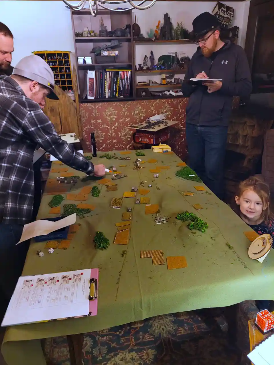 Three adults and a young girl are gathered around a large table covered with a green cloth, playing a miniature wargame with…