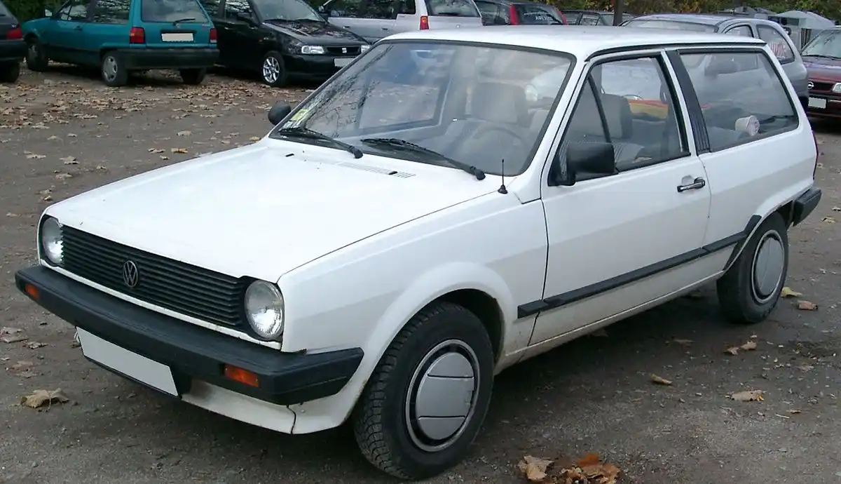 A white 1980s Volkswagen Golf Mk1 hatchback is parked in a lot with fallen leaves, showing its classic boxy design, black…