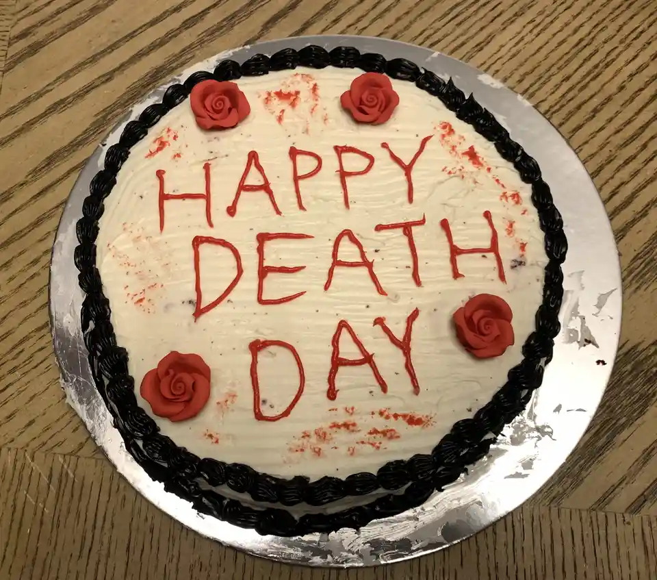 A round white-frosted cake with black piped icing border sits on a silver cake board atop a wooden table. Four red fondant…