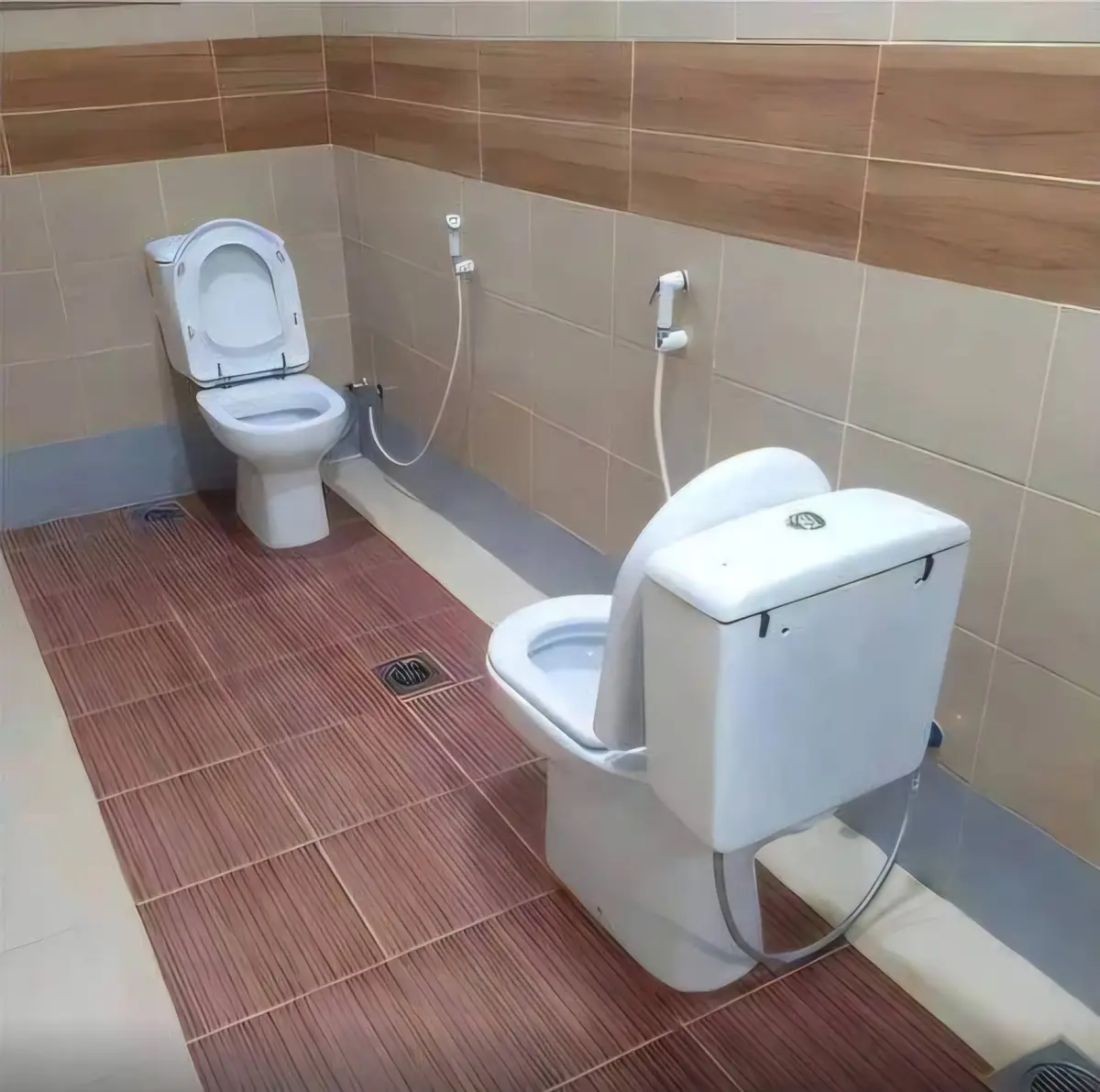A bathroom with two white toilets side by side on a reddish-brown tiled floor, walls are tiled with beige and brown…