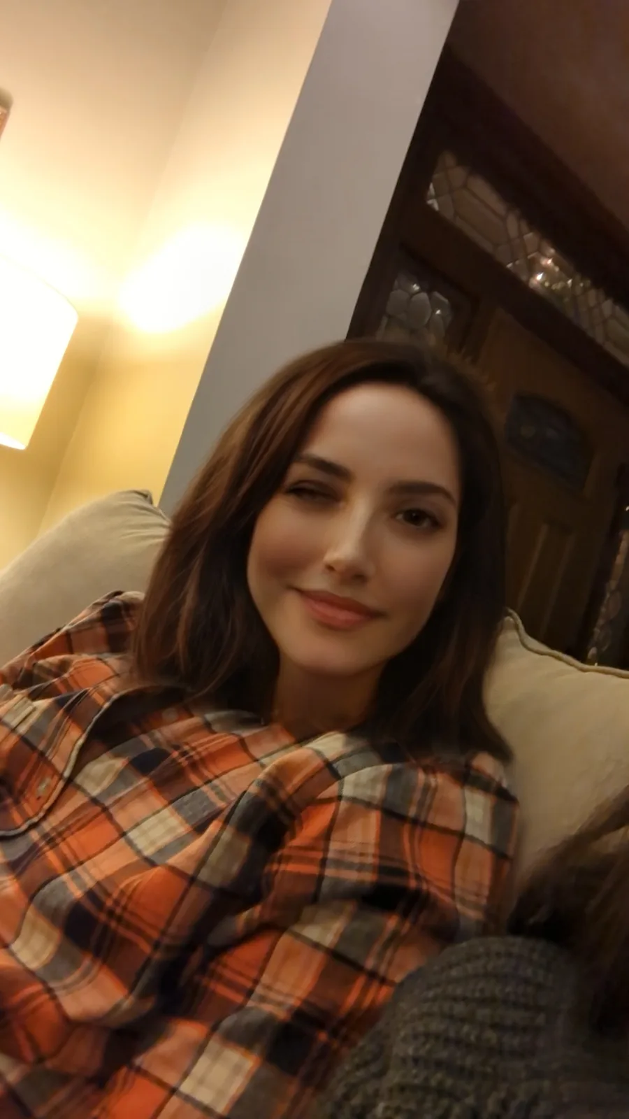 A woman with brown hair is lying on a couch wearing an orange, black, and beige plaid shirt, smiling slightly with one eye…
