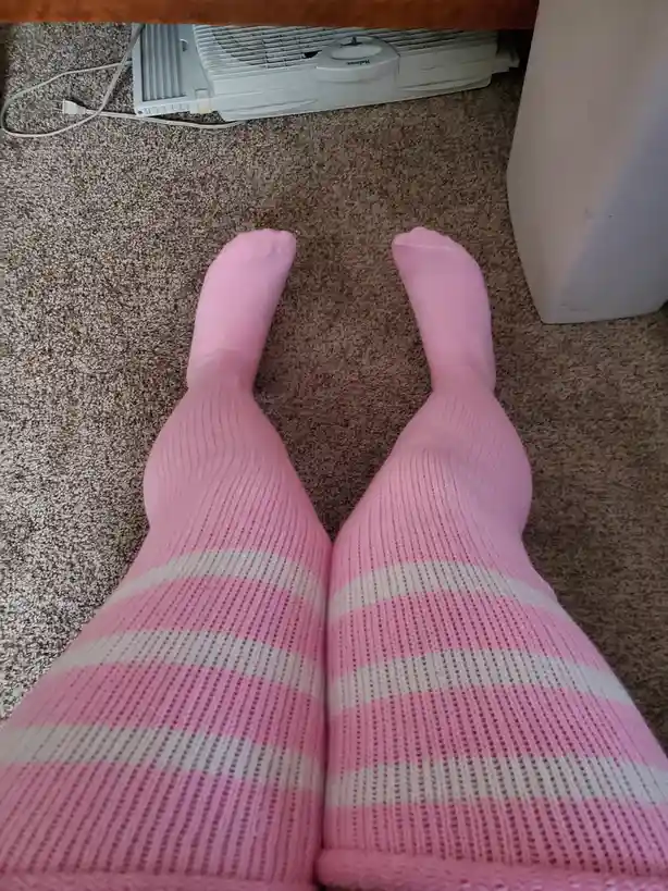 A top-down view shows a person's legs wearing pink, ribbed, knee-high socks with white horizontal stripes, resting on a…