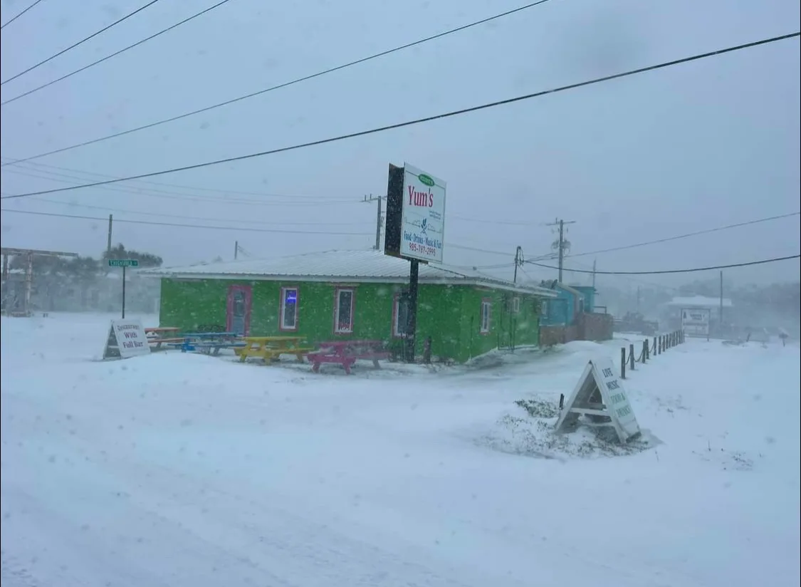 A bright green restaurant named 'Yum's' is surrounded by heavy snowfall, with picnic tables and a sign visible in the…