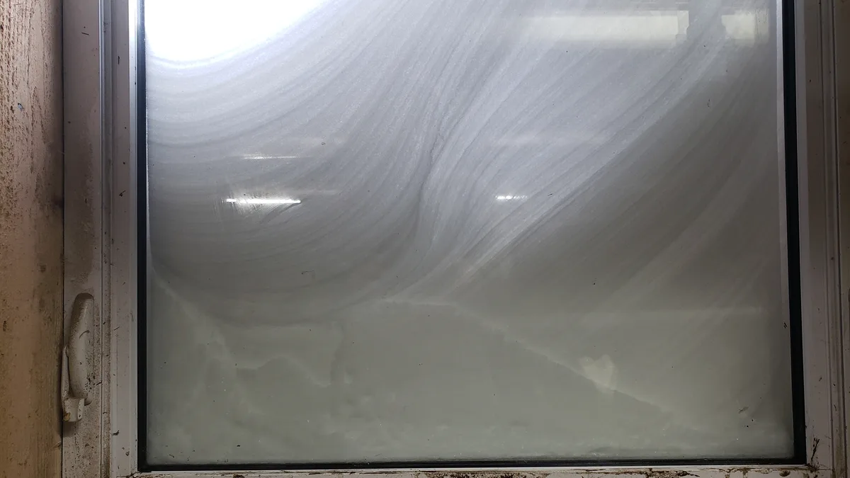 A close-up photo shows a dirty, grimy window with streaks of condensation or dirt across the glass, framed by a worn white…