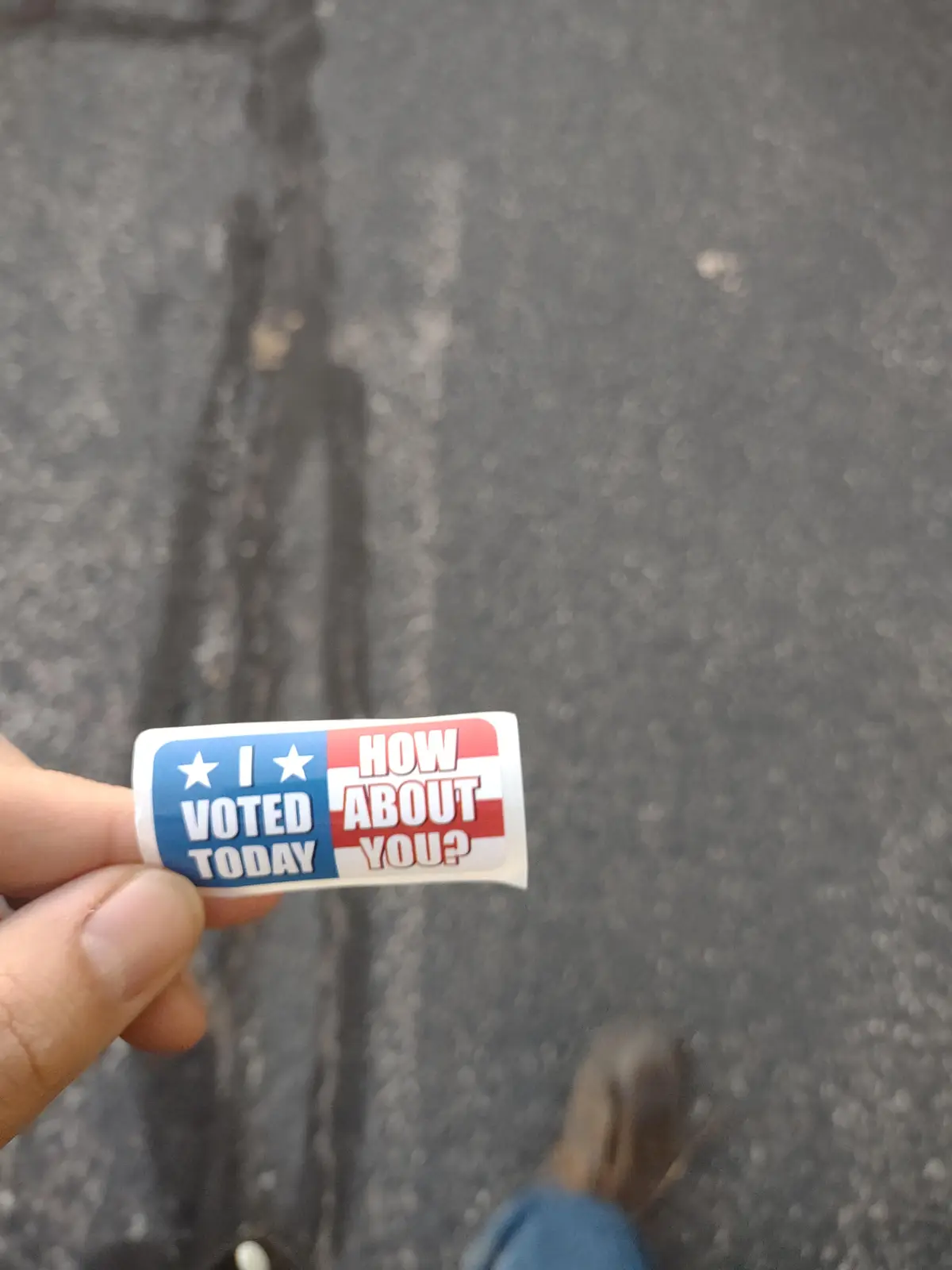 A hand holds a red, white, and blue sticker that reads 'I VOTED TODAY HOW ABOUT YOU?' against a gray asphalt background. The…