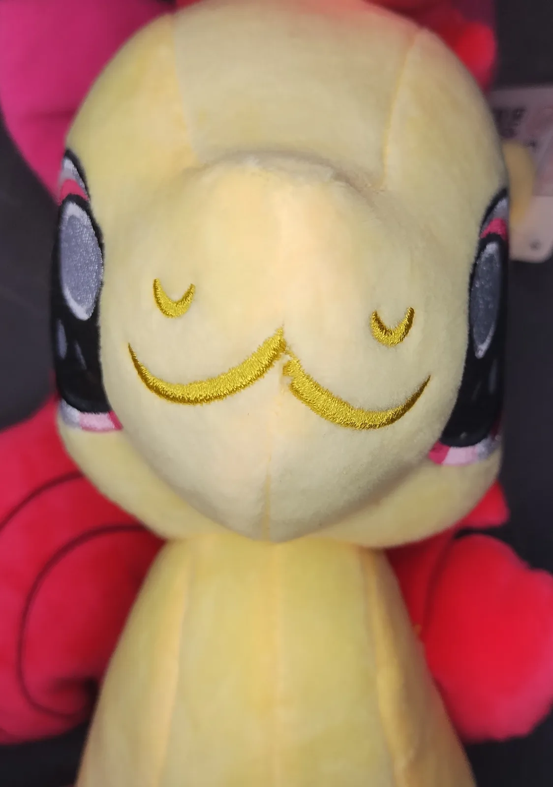 A close-up of a yellow plush toy with closed eyes and a wide, stitched smile, resembling a character from My Little Pony…