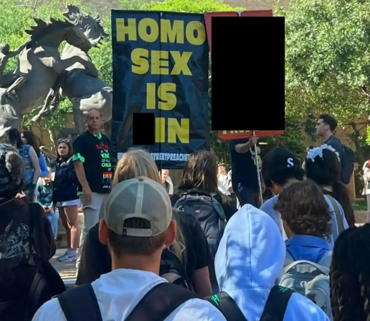 A crowd of people outdoors, some facing a large black banner with bold yellow text reading 'HOMO SEX IS IN', where the last…