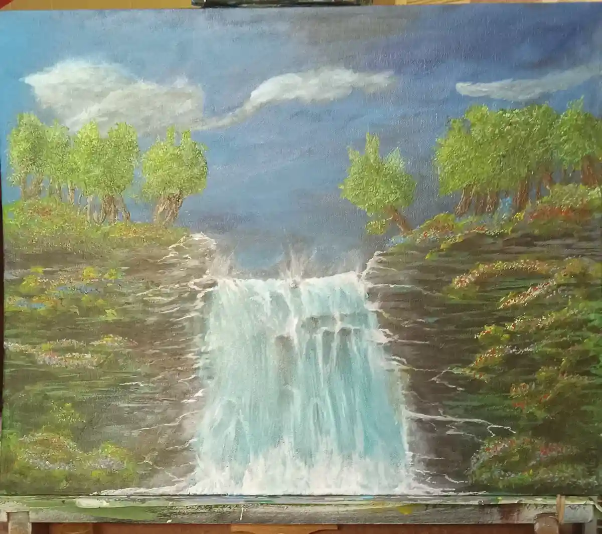 A painting of a waterfall surrounded by green trees and flowers under a blue sky with clouds, displayed on an easel. The…