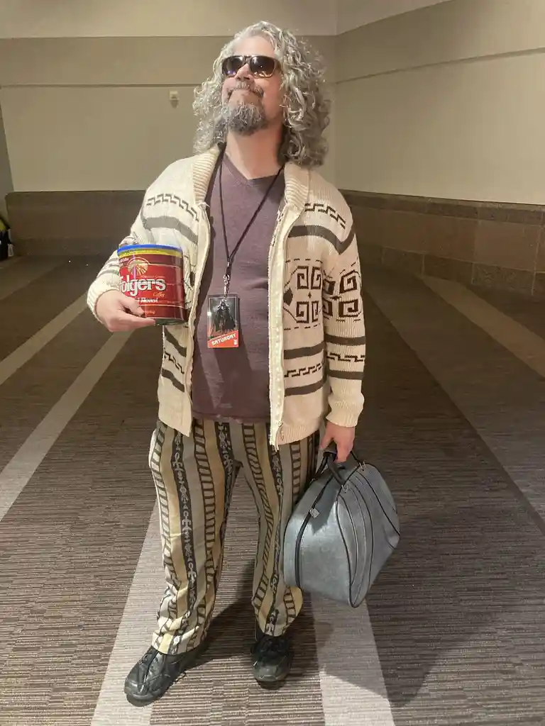 A man with long gray hair and sunglasses stands in a hallway, wearing a patterned beige cardigan, matching pants, and a…