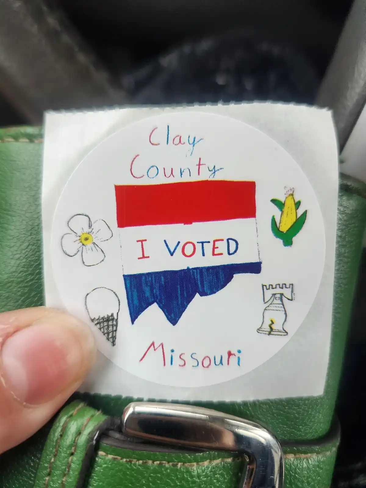 A hand holds a circular, hand-drawn 'I VOTED' sticker from Clay County, Missouri, featuring a red, white, and blue banner…