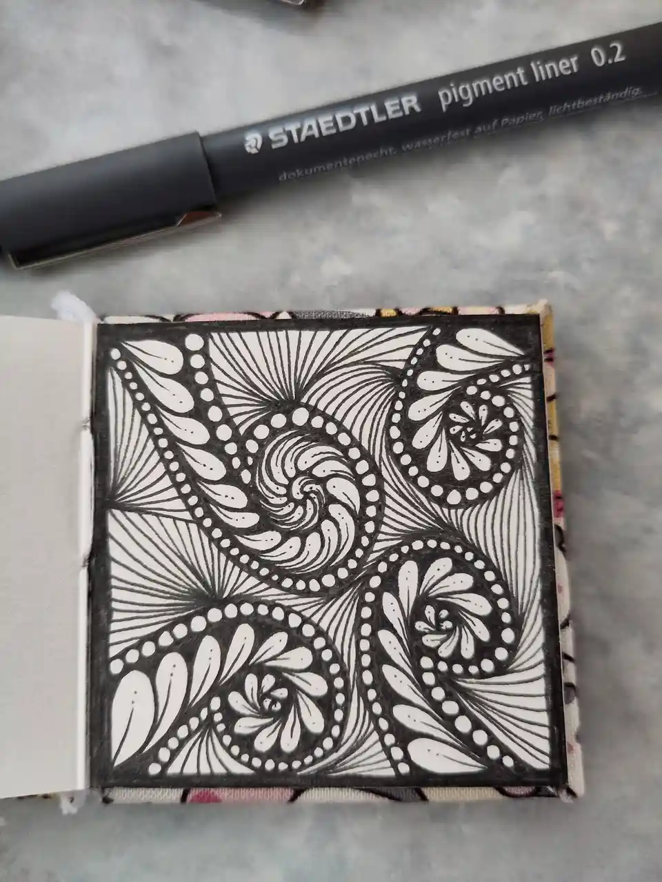 A close-up photo shows a small, open sketchbook with a highly detailed black-and-white zentangle-style drawing on the page…