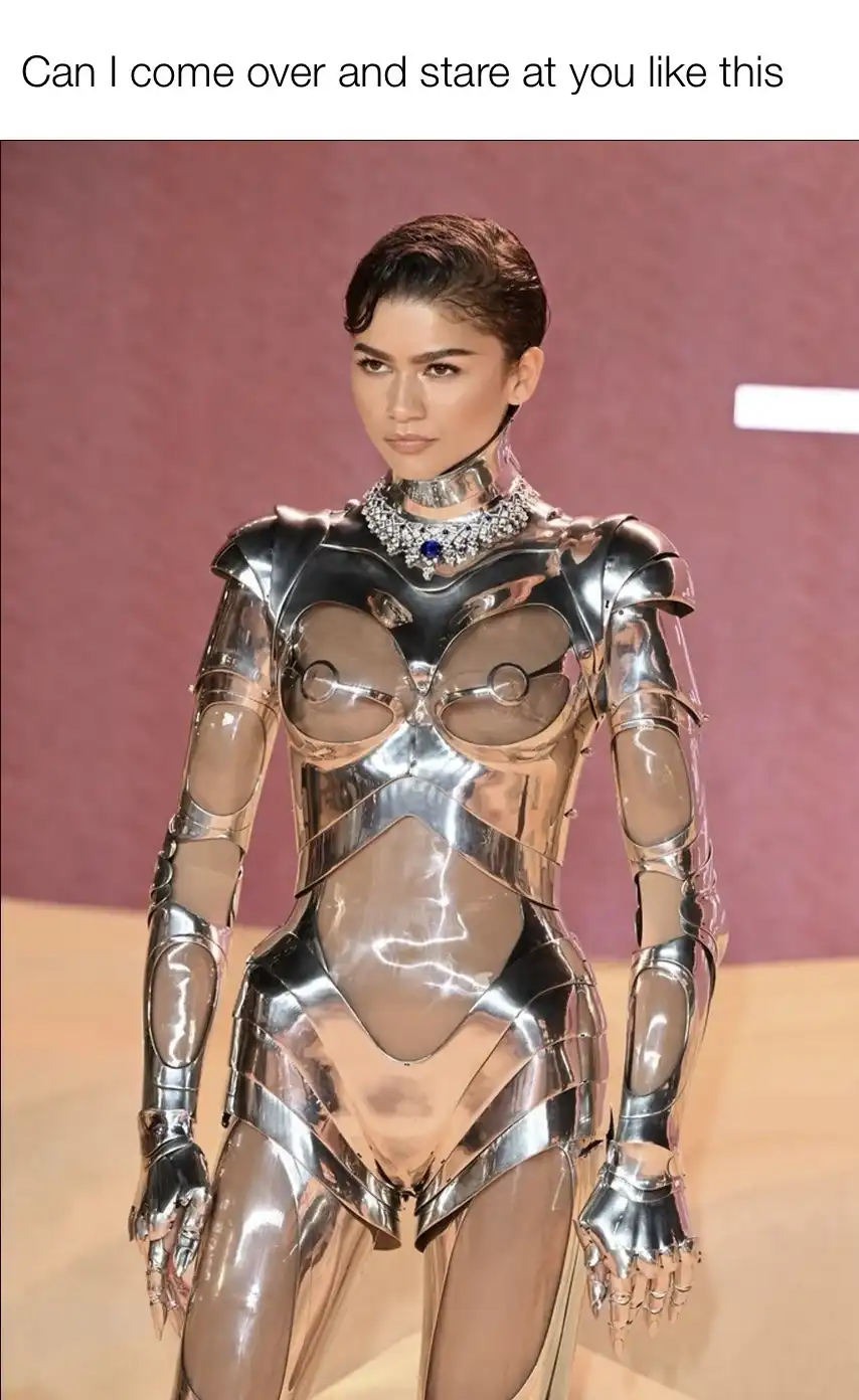 A woman with short dark hair stands in a shiny, metallic, segmented bodysuit with transparent panels revealing her skin…