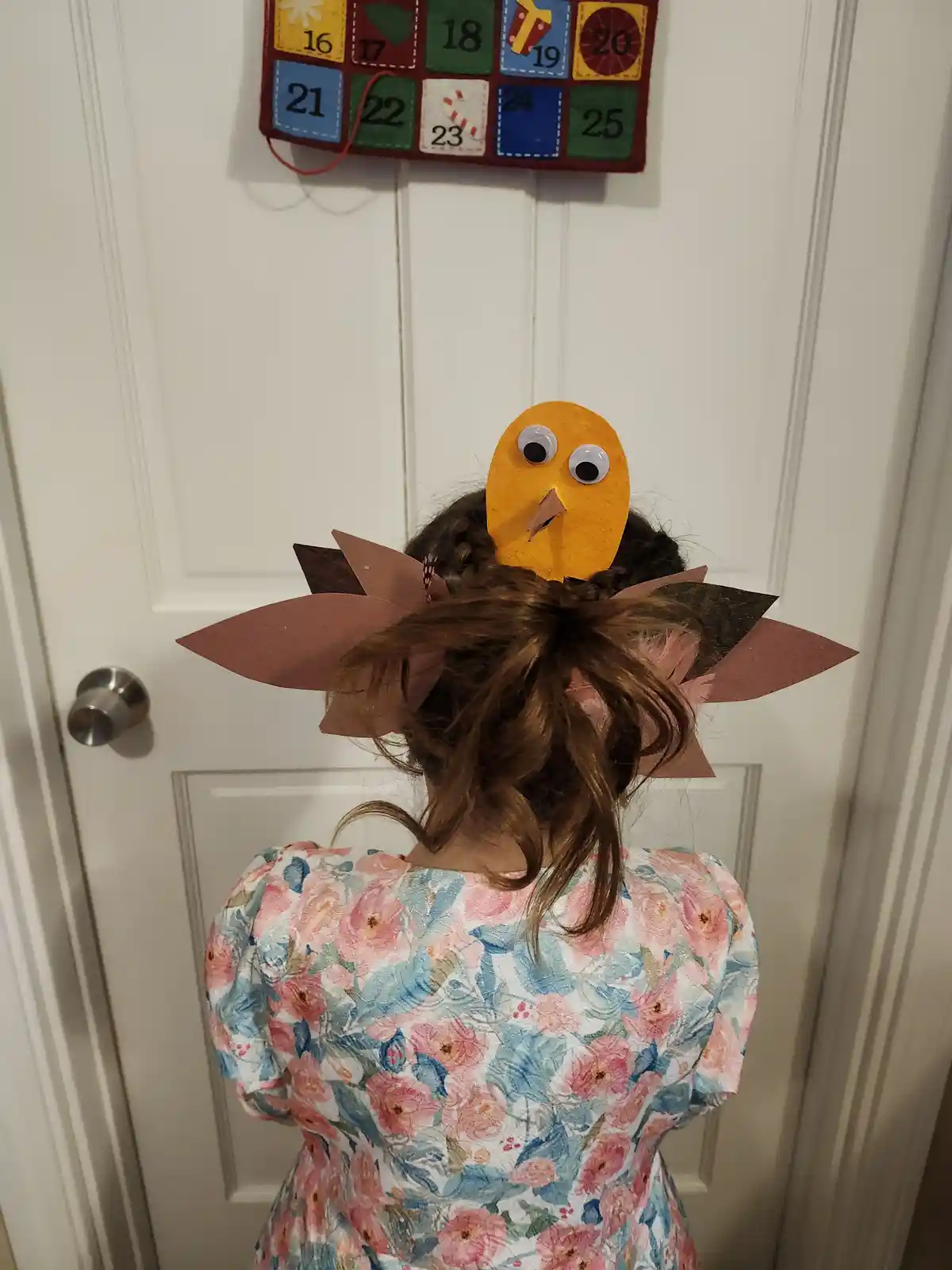 A child viewed from behind wears a handmade turkey headpiece made of orange felt, googly eyes, and brown paper feathers…
