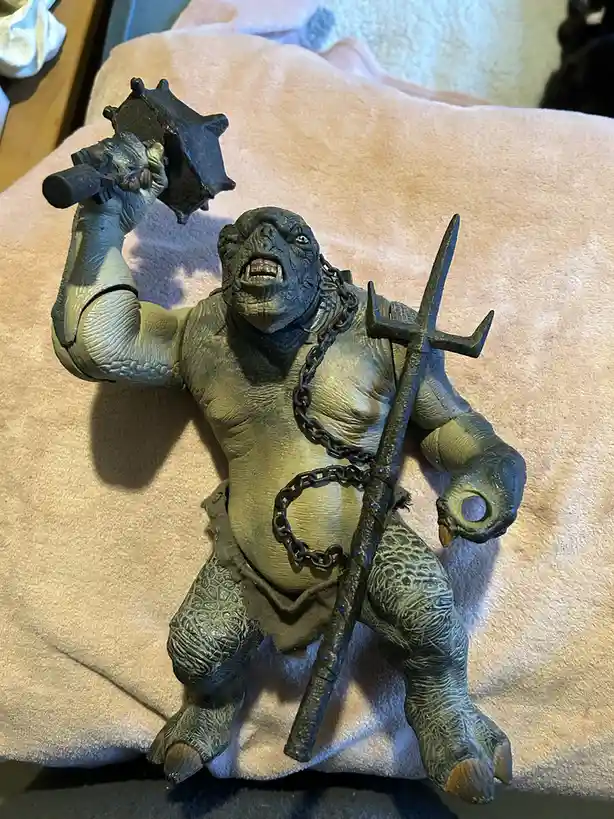 A green-skinned, muscular fantasy monster action figure with a chain around its neck and waist, holding a spiked mace in one…