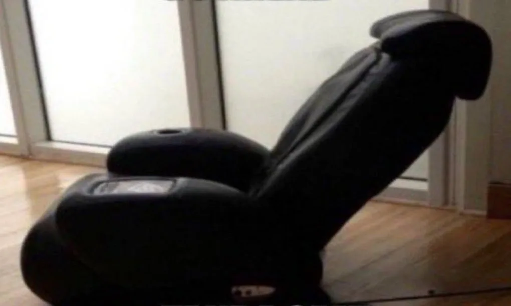 A black, reclining massage chair sits on a wooden floor in front of large windows, appearing unused and unremarkable. The…