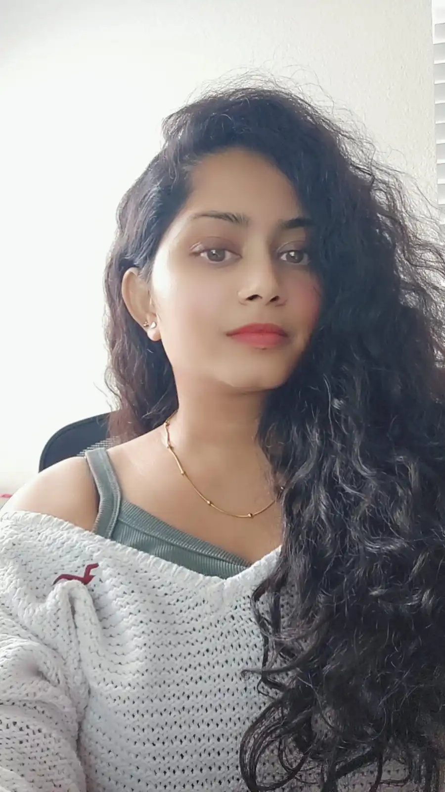 A young woman with long, dark curly hair looks directly at the camera with a neutral expression. She wears a white knitted…