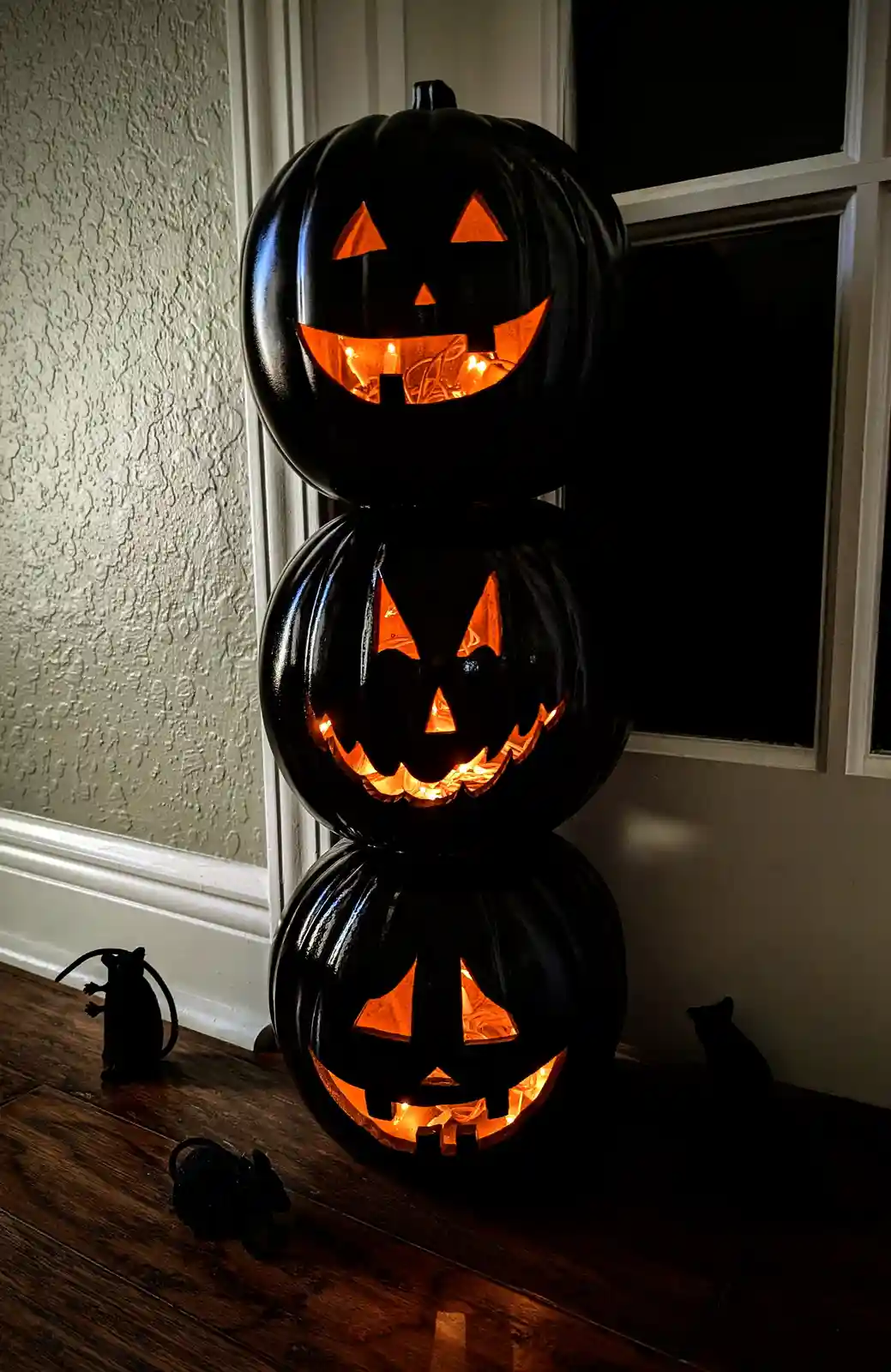 Three black, glowing jack-o'-lanterns are stacked vertically on a wooden floor near a window, each with a carved face…