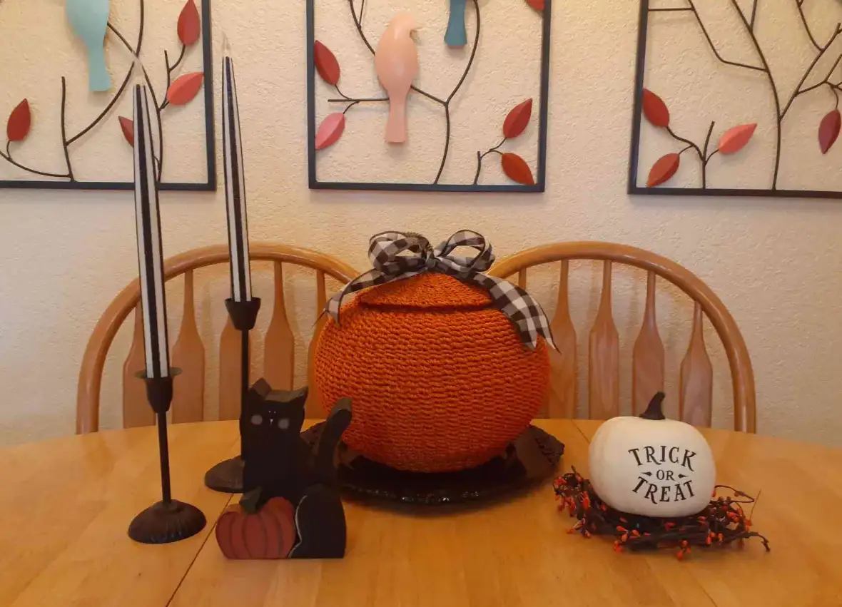 A wooden dining table displays Halloween decorations including a black cat figurine, two black candlesticks with white…
