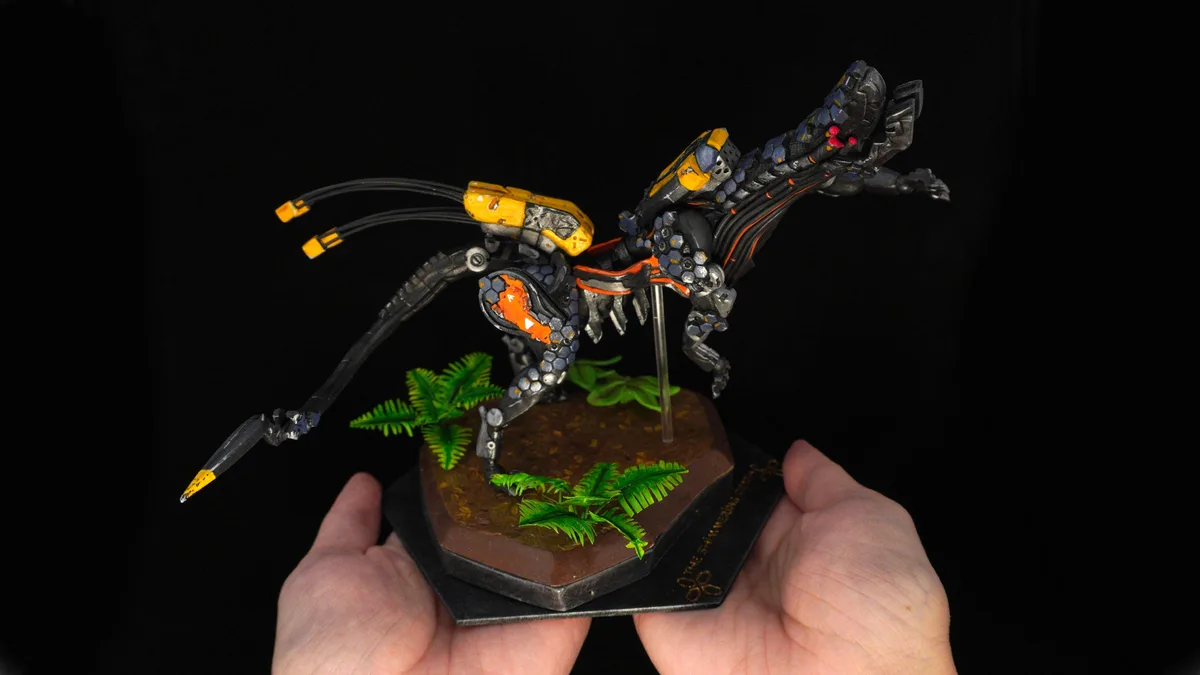Two hands hold a detailed, painted miniature of a biomechanical creature with black, orange, and yellow accents, standing on…