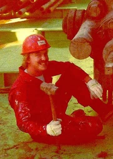 A man in a red hard hat and red coveralls sits on the ground holding a hammer, smiling. The image has a heavy red tint and…