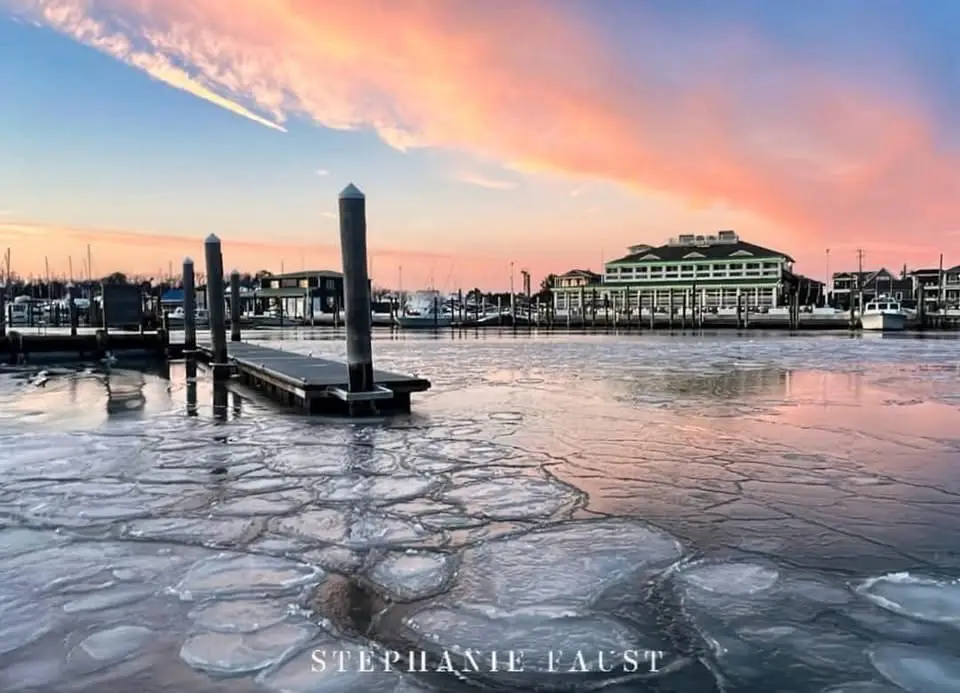A scenic photograph of a harbor at sunset, where the water is partially frozen with cracked ice floes in the foreground…