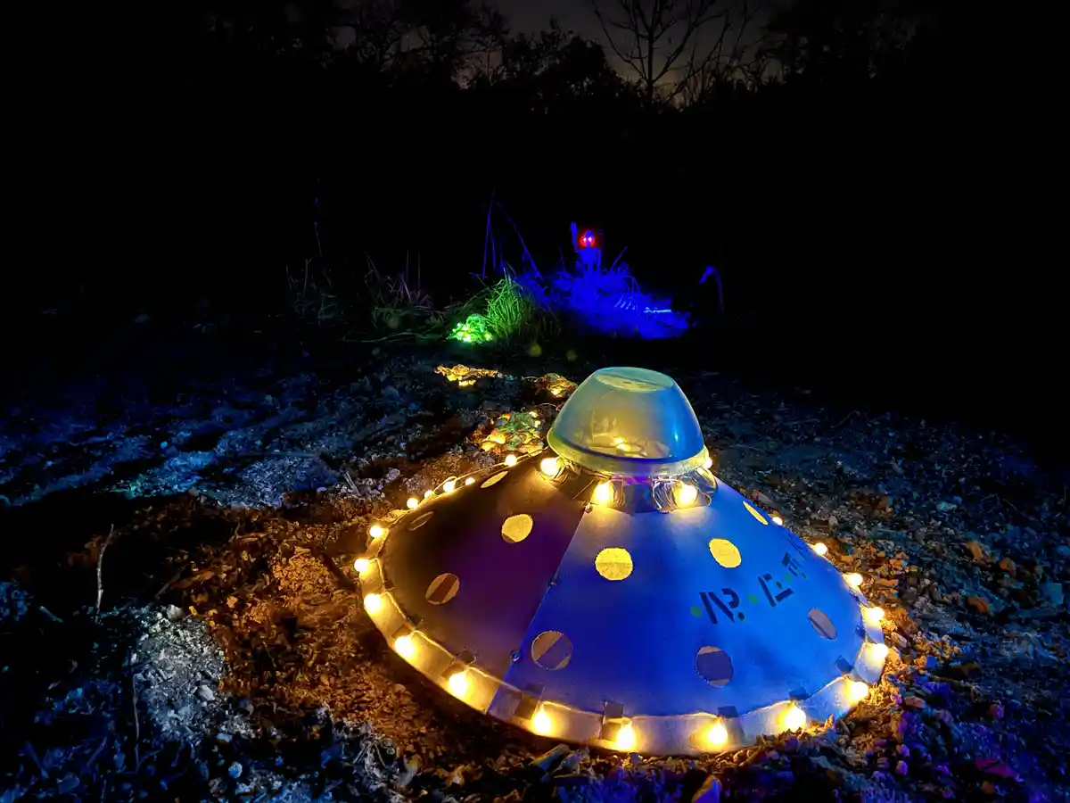A handmade, illuminated UFO decoration sits on dirt at night, surrounded by string lights, with another glowing alien figure…