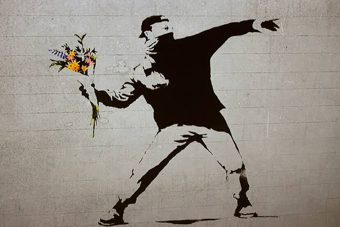 A stencil graffiti artwork depicts a masked figure in a dynamic throwing pose, holding a colorful bouquet of flowers in one…