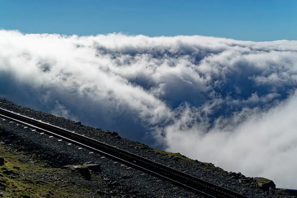 A scenic photograph shows a railway track ascending steeply on a rocky mountainside, with a vast sea of clouds below and a…