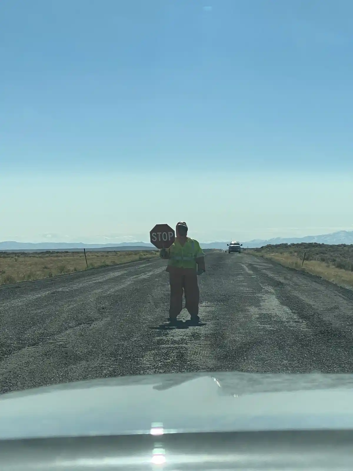 A person in a bright yellow safety vest and a face mask stands in the middle of a desolate, gravel road holding a red stop…