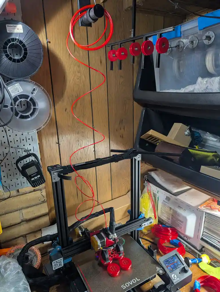 A 3d printer is shown actively printing small red circular objects, with a long red filament spool hanging from above and…