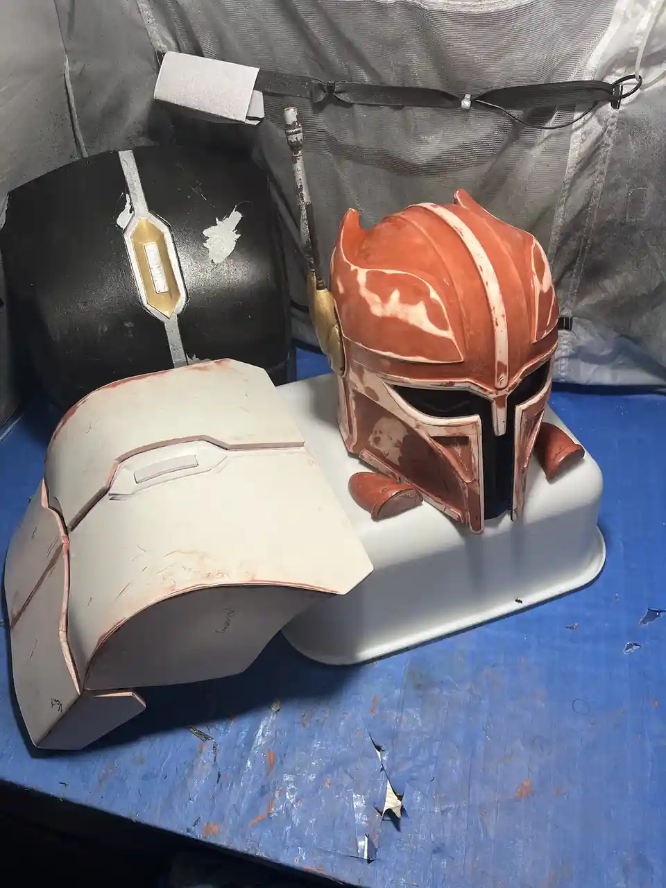 Three Mandalorian helmet molds sit on a blue workbench in a workshop; one is black with gold accents, one is orange with…