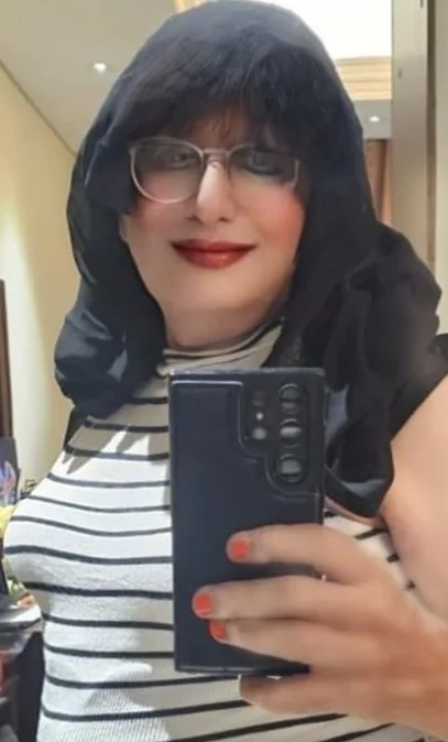 A woman with dark hair, glasses, and red lipstick takes a mirror selfie holding a black smartphone with a prominent camera…
