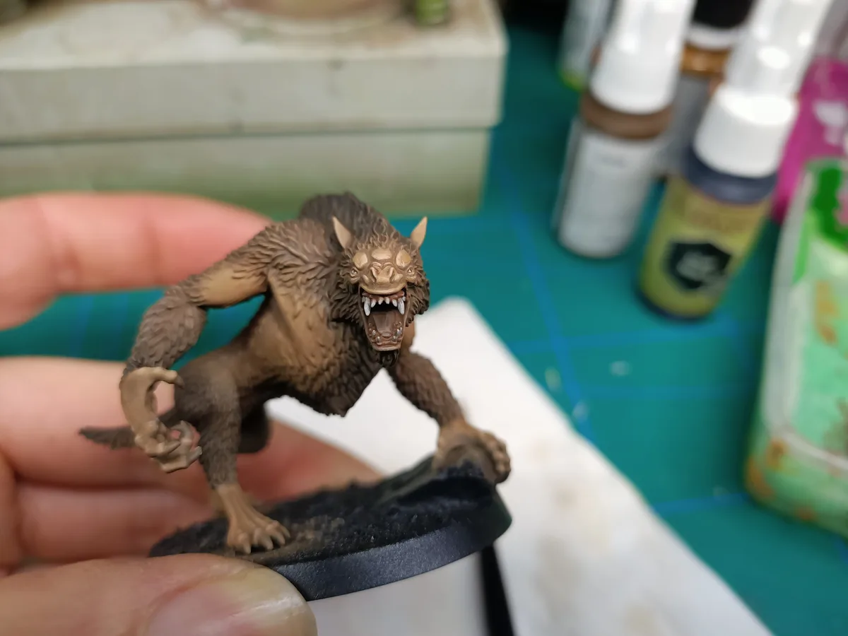A hand holds a detailed, brown-painted miniature of a snarling, furry werewolf-like creature with sharp teeth and claws…