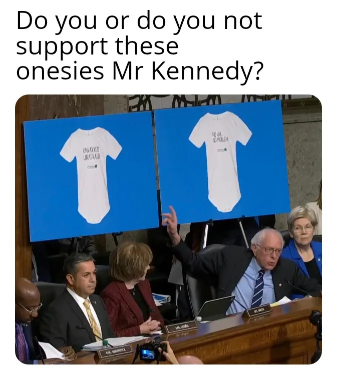 A photo shows Senator Bernie Sanders pointing at two large blue boards displaying white baby onesies. The onesie on the left…