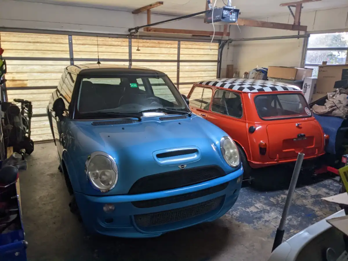 Two Mini Coopers are parked side-by-side in a cluttered garage; one is a modern blue car in the foreground, and the other is…