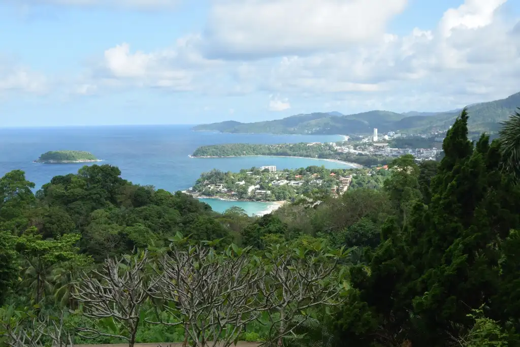 A scenic tropical landscape with lush green trees in the foreground, a turquoise bay, a coastal town, and distant hills…
