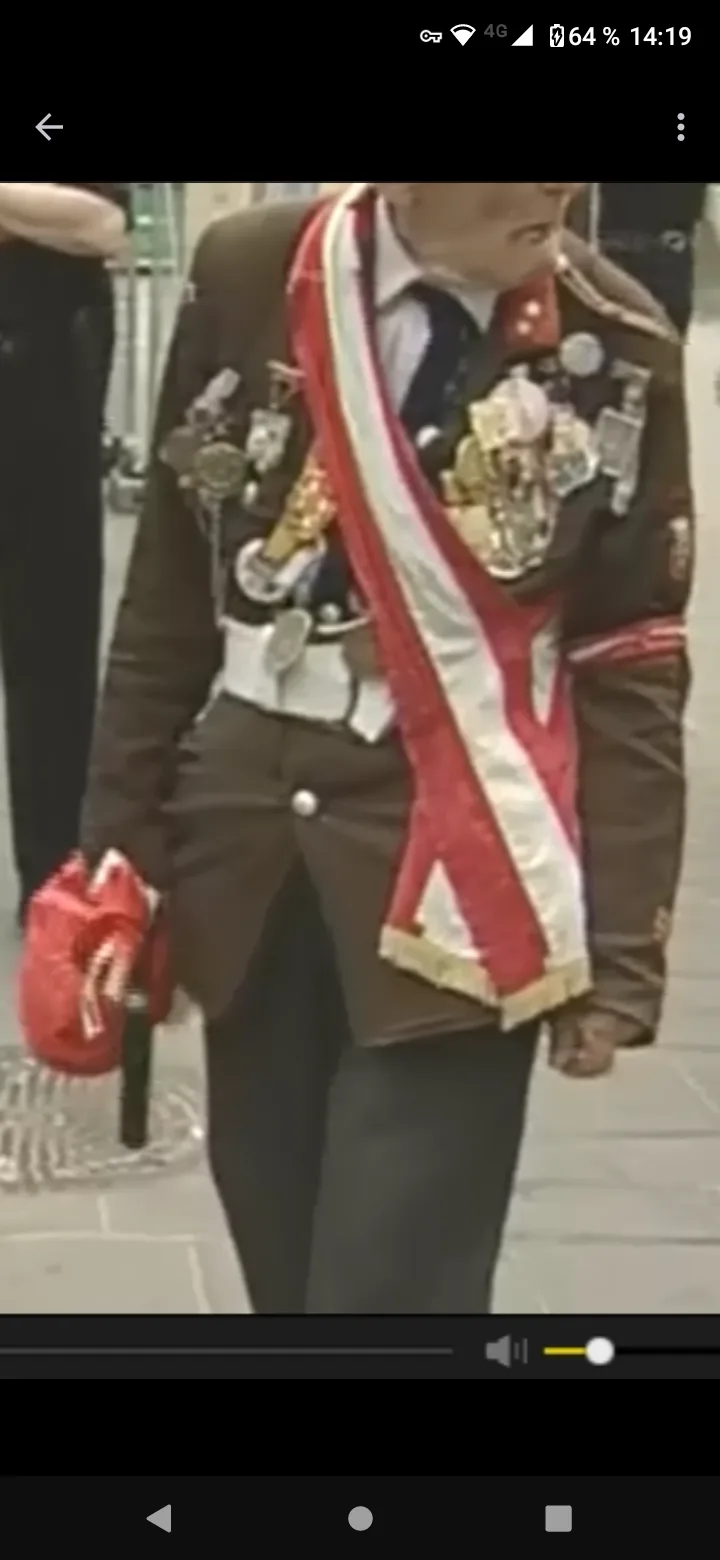 A person in a formal brown uniform adorned with numerous medals and a red-and-white sash walks while holding a red bag. The…