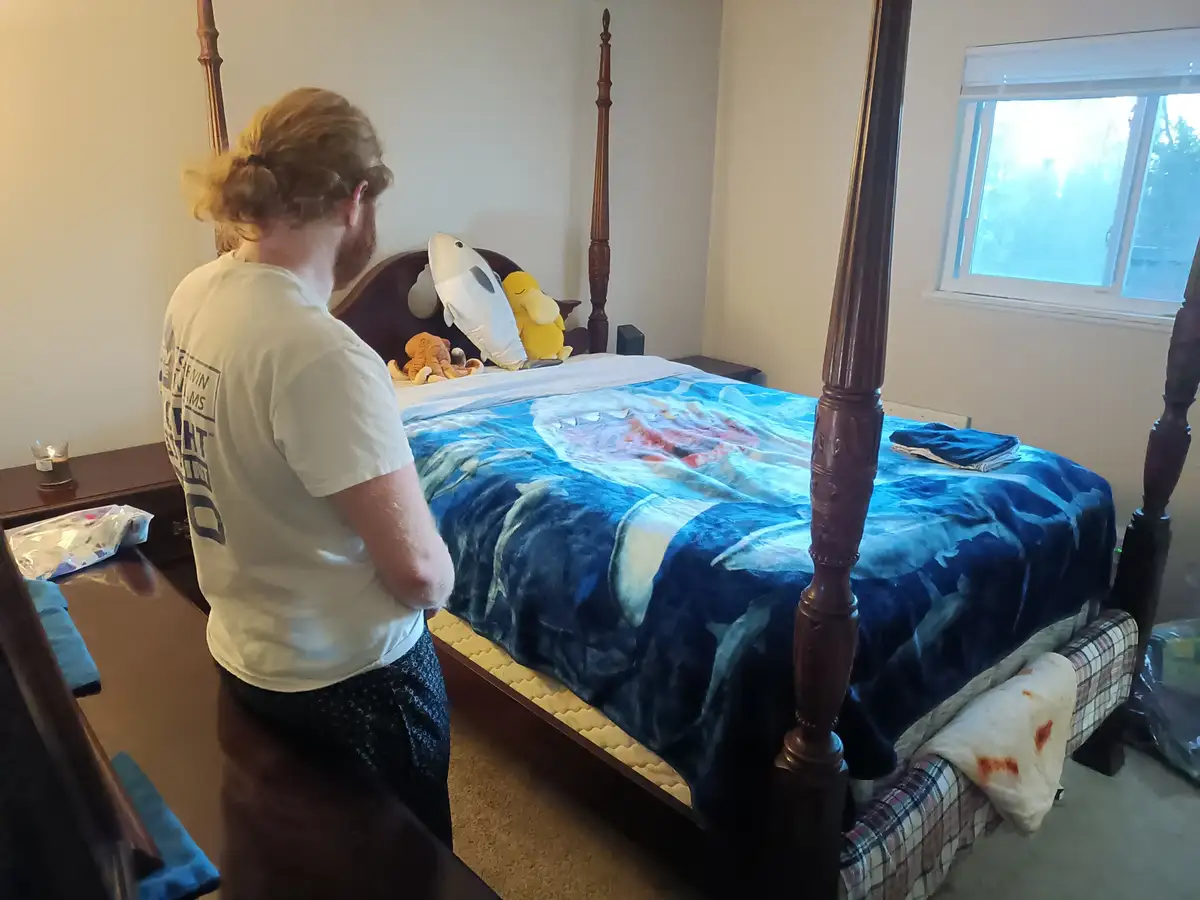 A person with long hair tied back stands in a bedroom, facing a four-poster bed with a blue dolphin-themed blanket. Several…