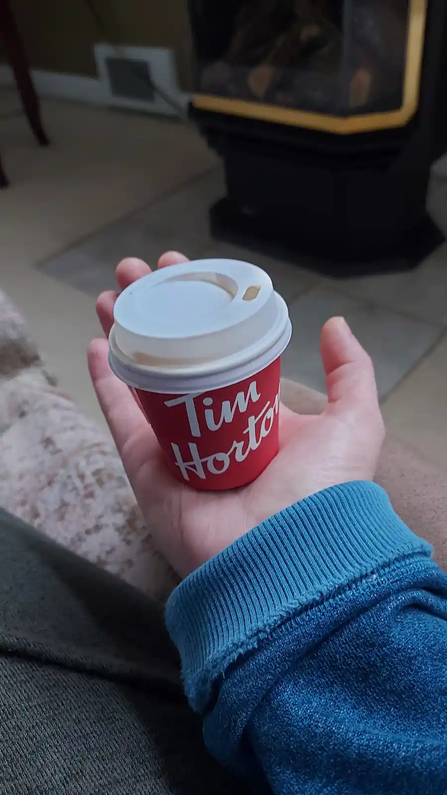 A person's hand holds a red Tim Hortons coffee cup with a white lid, wearing a blue sweater, in front of a fireplace in a…