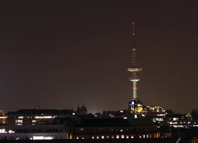A nighttime cityscape photograph showing a tall, illuminated tower with red lights against a dark sky, surrounded by lit…