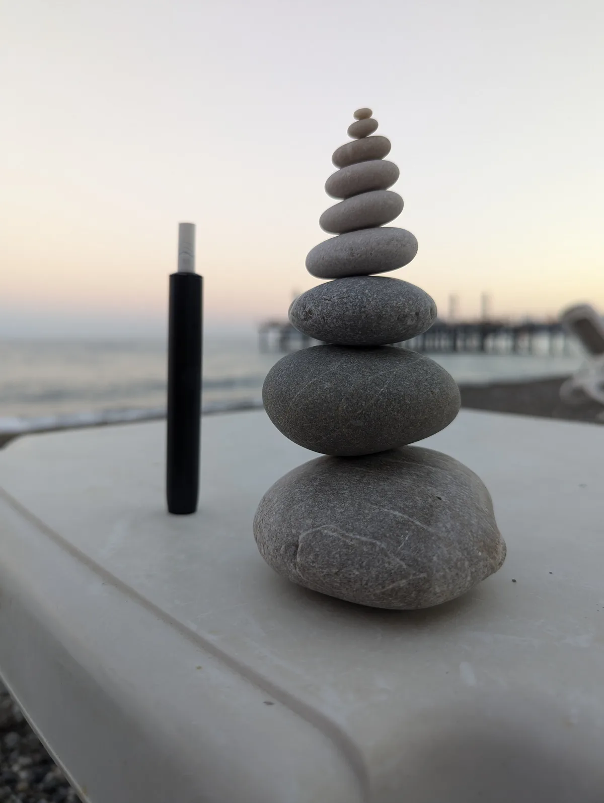 A tall, perfectly balanced stack of smooth, rounded stones sits on a white surface near a beach at dusk, next to a black…