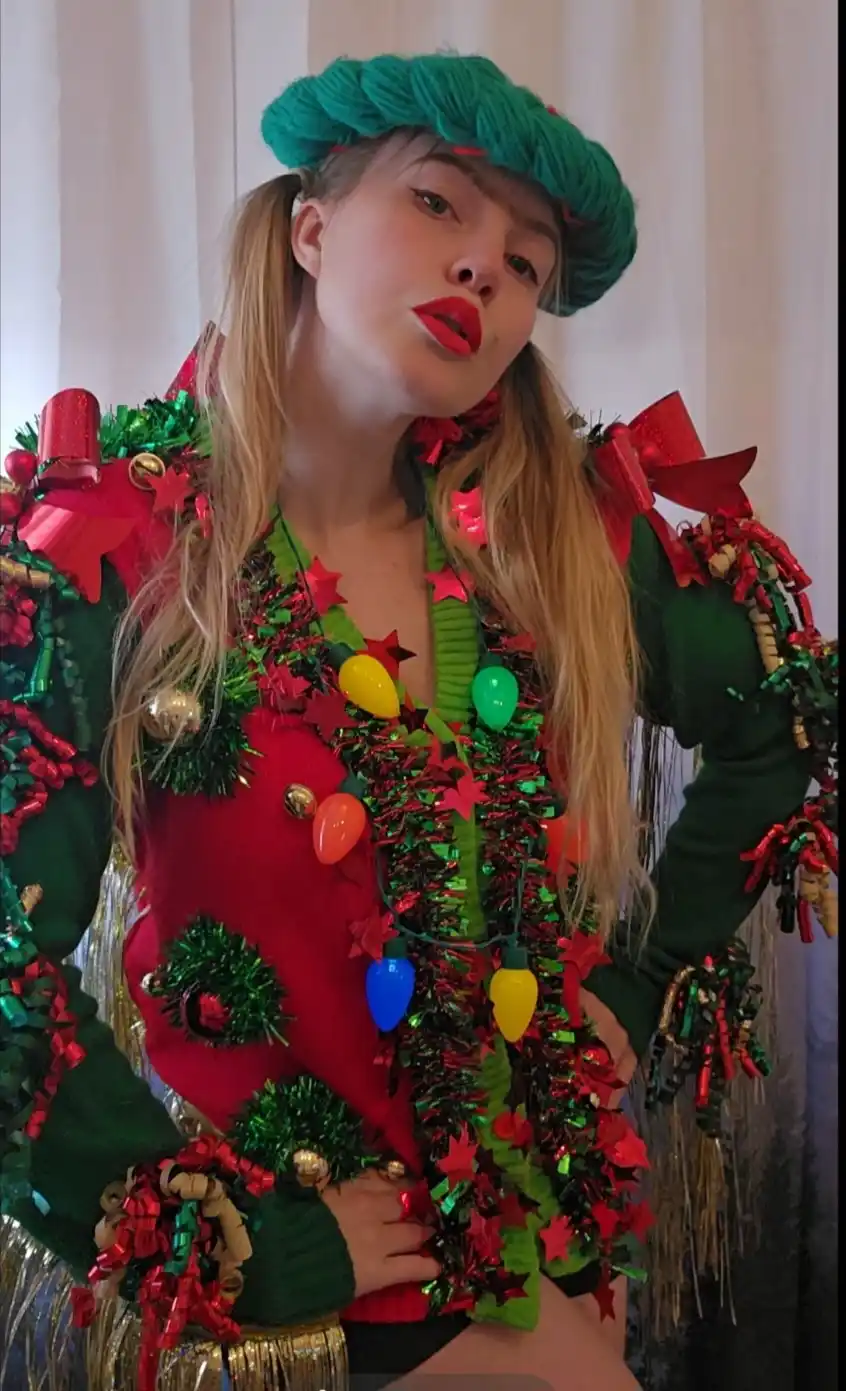 A woman poses in a heavily decorated holiday outfit, wearing a green knit beret, a red and green sweater covered in tinsel…