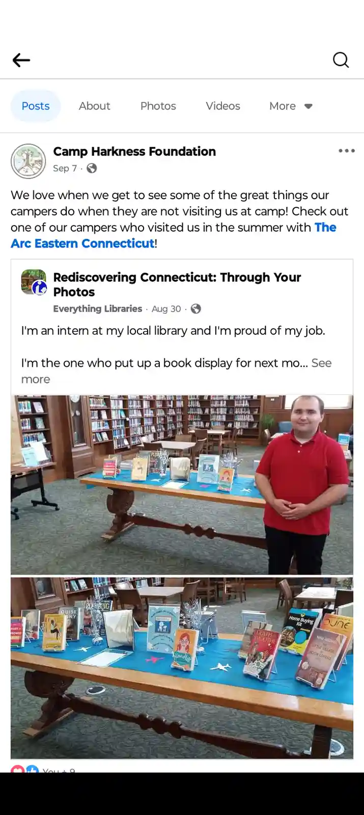 A man in a red polo shirt stands proudly beside a long wooden table covered with a blue cloth, displaying various books in a…
