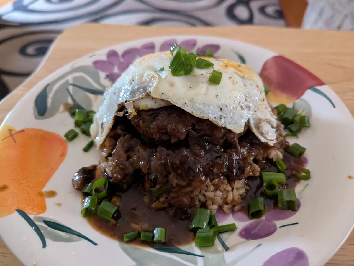 A close-up photo of a plated meal featuring a hamburger patty covered in a dark sauce and rice, topped with a fried egg and…